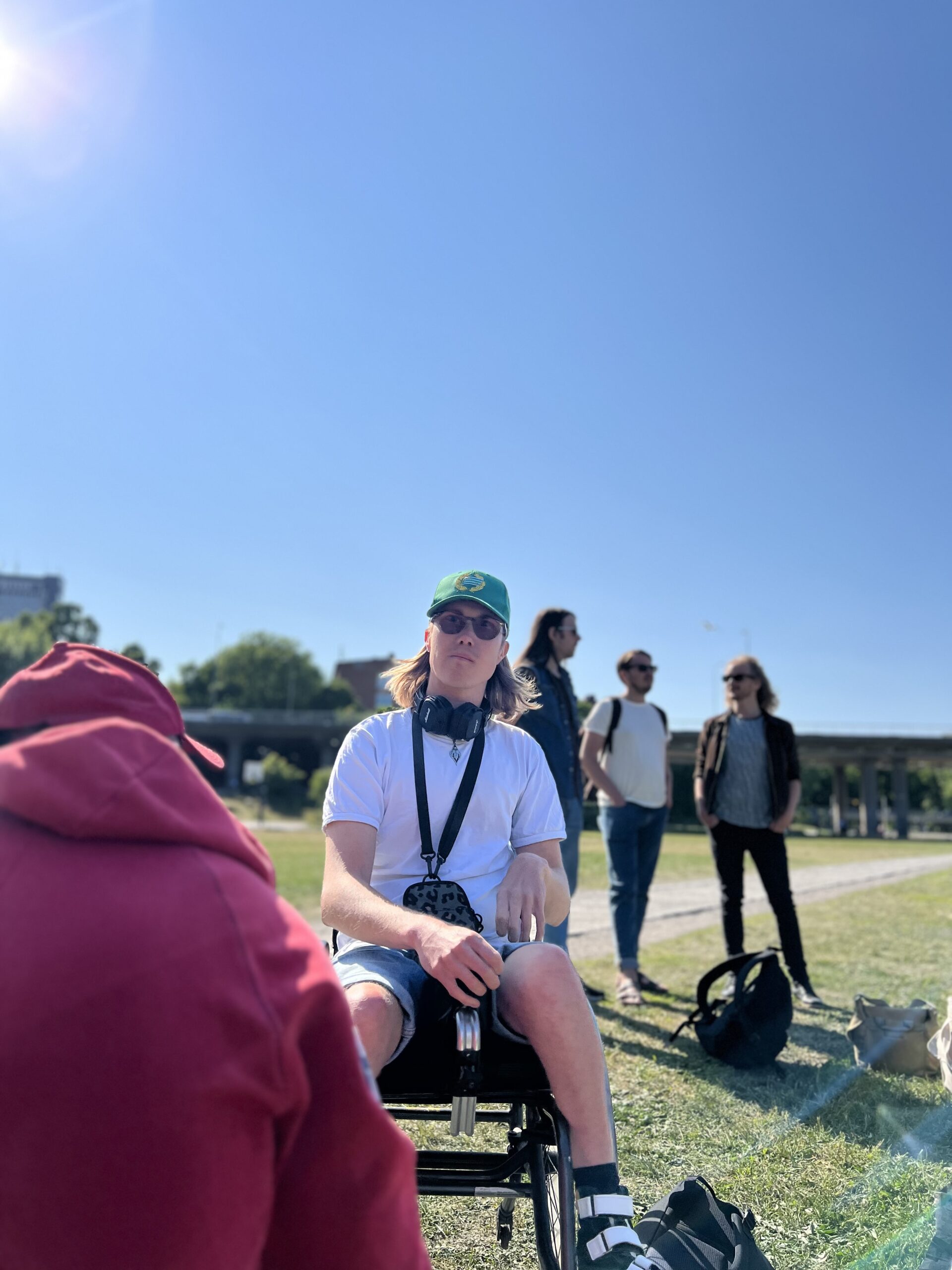 Ung person i grön keps och solglasögon sitter i fokus med hörlurar runt halsen, medan tre andra personer står suddigt i bakgrunden i en park under klarblå himmel.